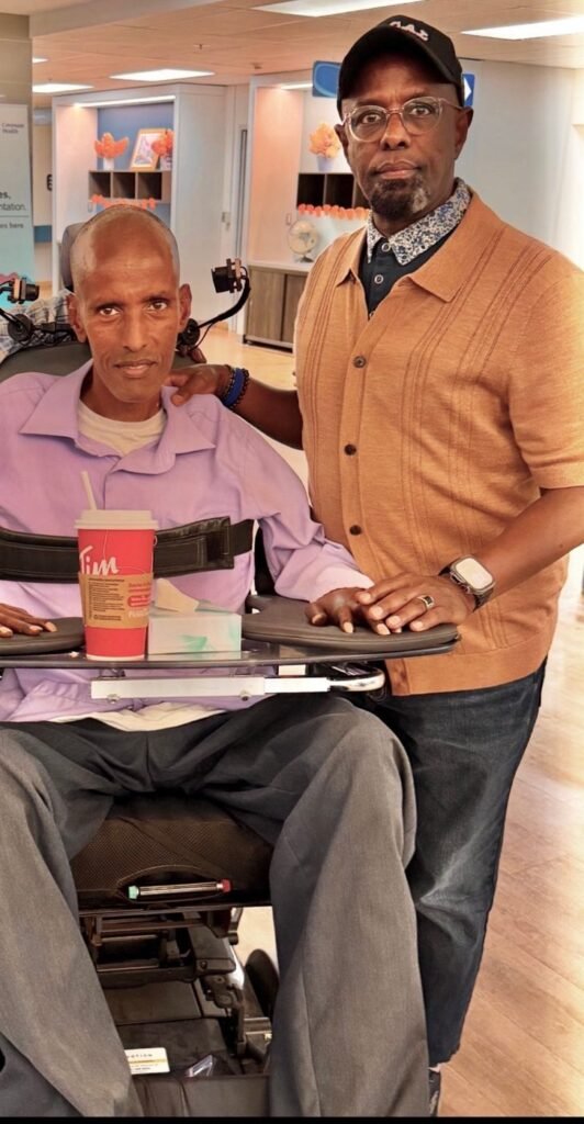 Chairman Ibrahim Shurie standing beside a community member using a power wheelchair in a hospital lounge