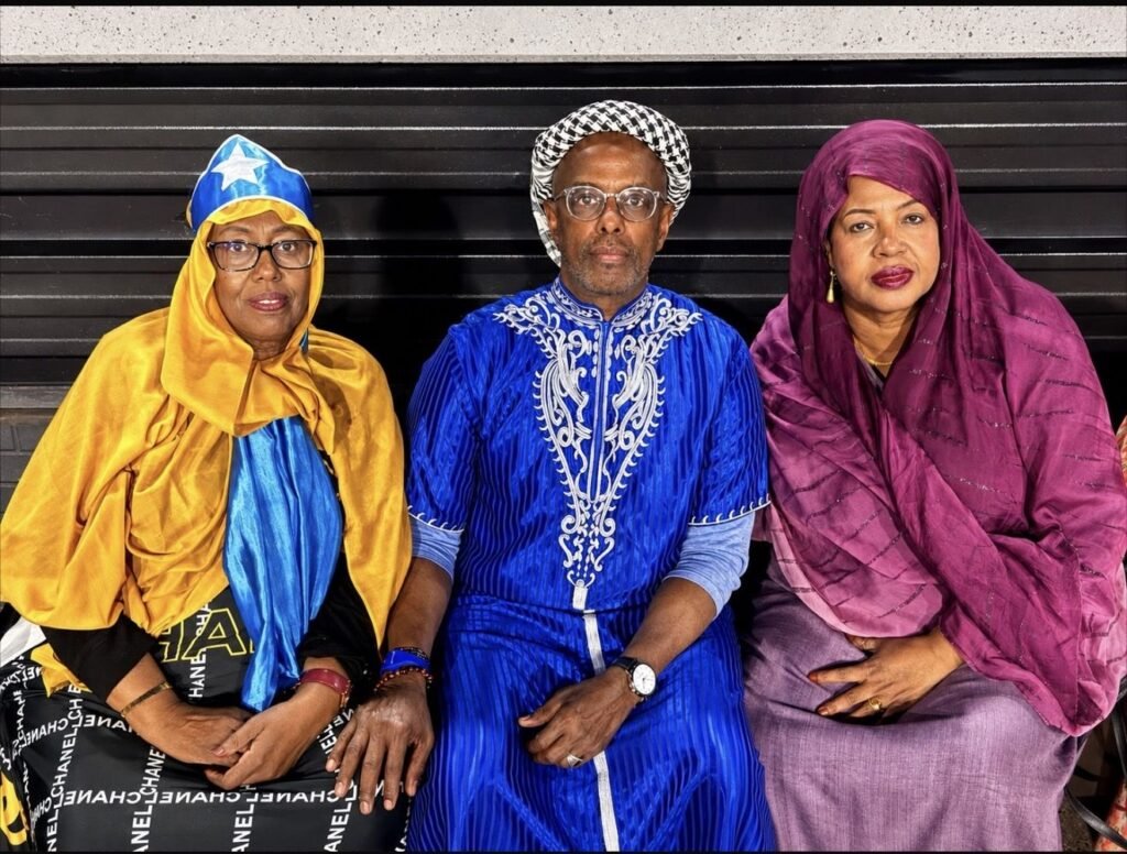 Three Somali community elders in colourful traditional dress seated together, one wearing Somali flag colours