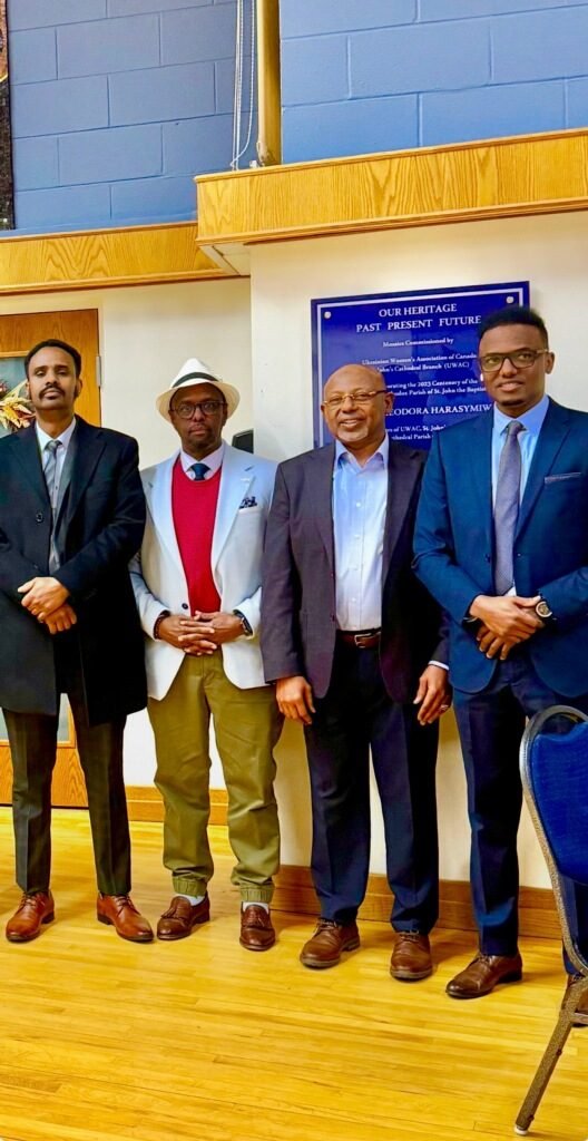 Four community leaders standing together in a community hall beside a heritage plaque